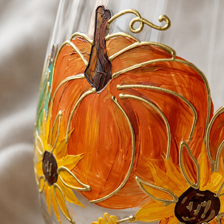 A clear glass wine glass decorated with a hand-painted autumn design featuring orange pumpkins, yellow sunflowers, and gold leaf-like lines, resting on a beige fabric surface.