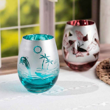 Two decorative stemless wine glasses on a white tablecloth in front of a softly blurred window. The front glass is frosted white with turquoise blue lower section and tropical etched designs including a dolphin, palm tree, waves, sun, and a shark.