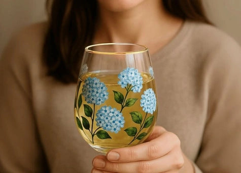 A person in a beige sweater holds a stemless wine glass with a gold rim and hand-painted blue flowers and green leaves, filled with a pale yellow drink.