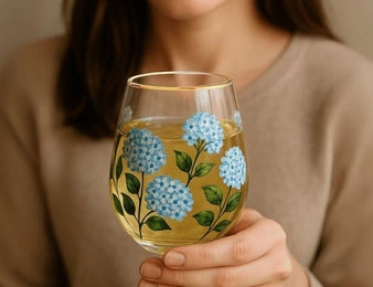 A person in a beige sweater holds a stemless wine glass decorated with painted blue hydrangeas and green leaves, filled with a light golden beverage.