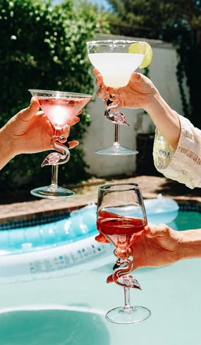 Three hands holding cocktail glasses with sculpted flamingo-shaped stems, toasting by a swimming pool outdoors. The glasses contain pink, white, and red cocktails, with greenery and a pool in the background.
