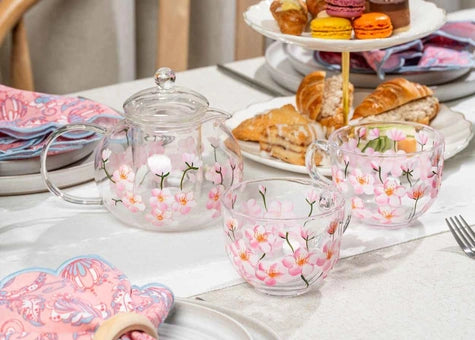 A clear glass teapot and two matching teacups decorated with pink cherry blossom flowers, arranged on a white tablecloth with pastries and napkins in the background.