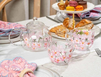 A clear glass teapot and matching cups decorated with pink cherry blossom flowers, arranged on a white tablecloth with pastries and napkins in the background.