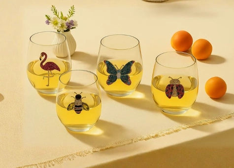 Four clear stemless wine glasses filled with yellow liquid, each decorated with a different insect or bird illustration, arranged on a beige surface with decorative oranges and a small vase of flowers.