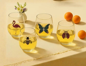 Four clear stemless wine glasses filled with yellow liquid and decorated with animal illustrations, arranged on a beige surface with oranges and a small vase of flowers in the background.