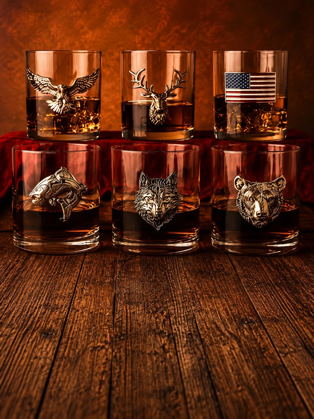 Six clear whiskey glasses arranged on a wooden surface against a warm brown background. Each glass contains amber liquid and has a different metallic animal or flag emblem on the front: eagle, deer, American flag, shark, wolf, and bear.