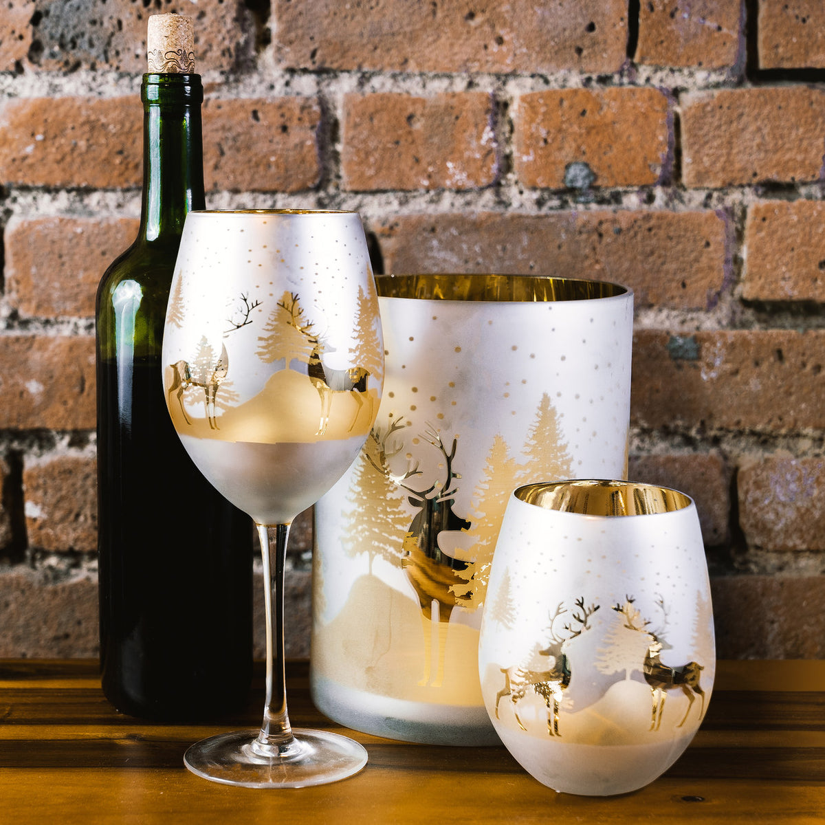Gold and silver deer candle holder with matching wine glass and stemless tumbler on a wooden table against a brick wall