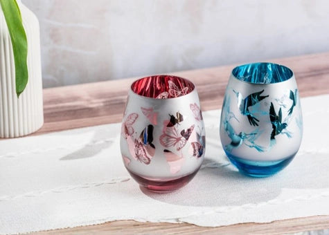 Two decorative stemless wine glasses with butterfly and bird motifs, one in pink and one in blue, set on a white table runner.