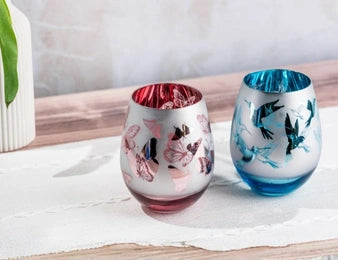 Two decorative stemless wine glasses with butterfly and bird motifs, one in pink and one in blue, set on a white cloth on a wooden surface.