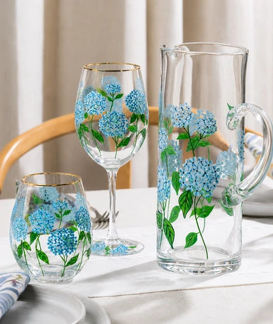 A clear glass dining set with blue hydrangea floral prints and gold trim, including a pitcher, a wine glass, and a tumbler, arranged on a white tablecloth.