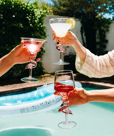 Three hands holding cocktail glasses with flamingo-shaped stems and rims, clinking together near a swimming pool outdoors. The glasses contain pink, white, and red cocktails, with green foliage and a white wall in the background.