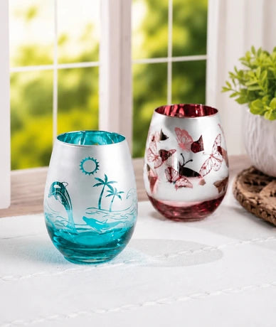Two decorative stemless wine glasses on a white table runner in front of a window. The left glass is frosted white with turquoise blue tropical ocean and dolphin designs.