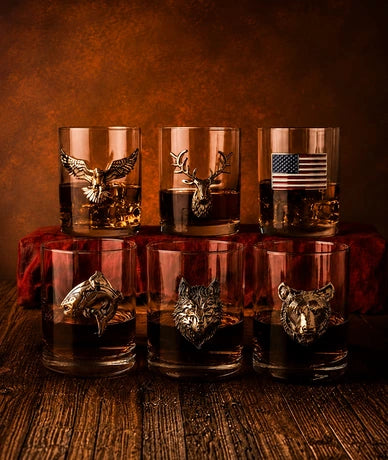 Six clear whiskey rocks glasses with metallic animal and flag engravings, arranged on a wooden surface against a warm brown background. The top row has three glasses with eagle, deer, and American flag designs, and the bottom row has three glasses with fish, wolf, and tiger designs.