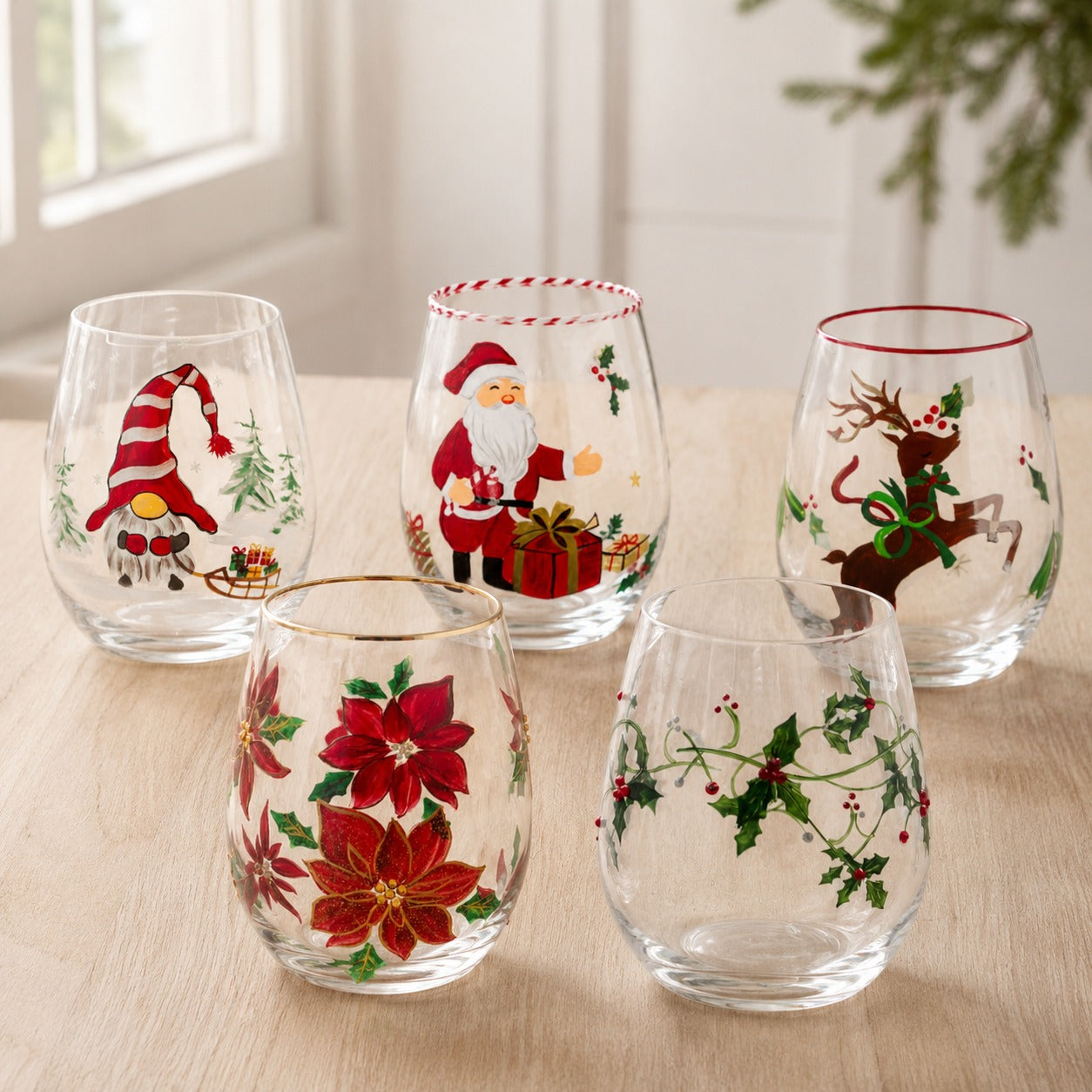 Collection of hand-painted Christmas stemless wine glasses on a sunlit table - gnome, Santa, reindeer, poinsettia and holly ivy designs