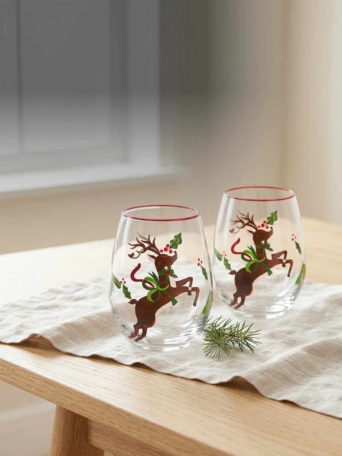 Both hand-painted reindeer stemless wine glasses together on a sunlit oak table