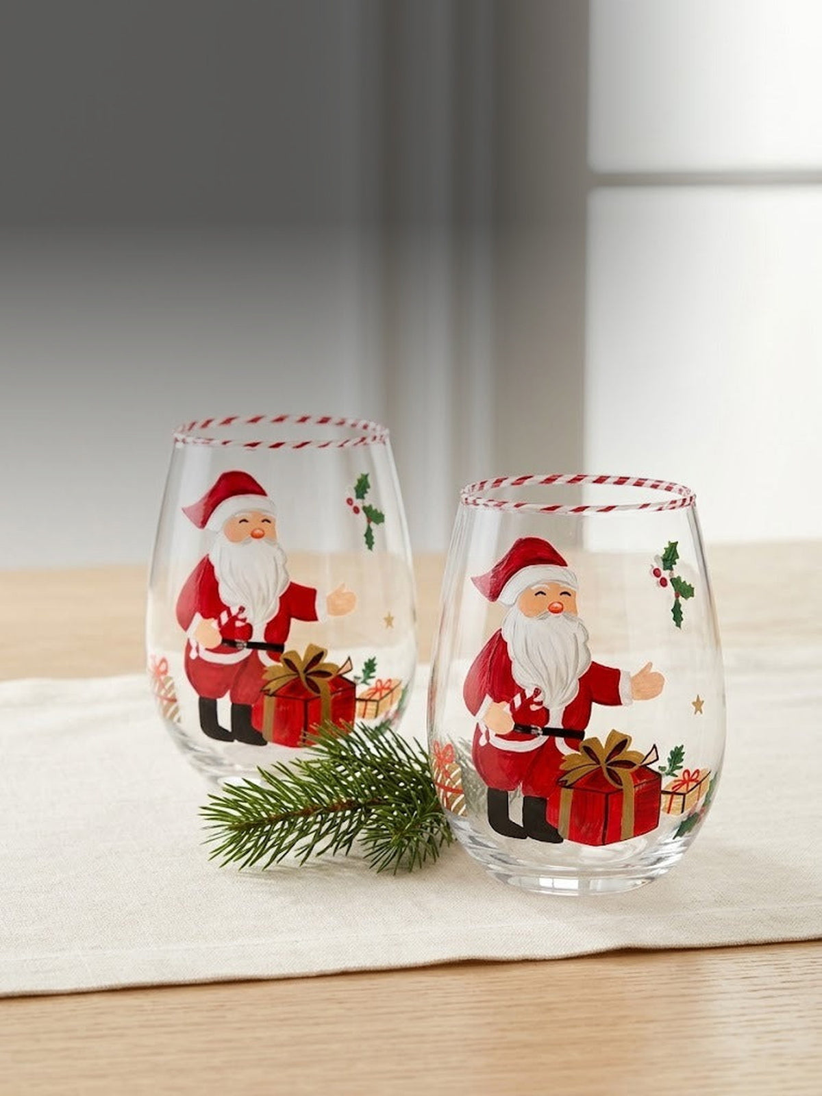 Both hand-painted Santa stemless wine glasses together on a sunlit oak table