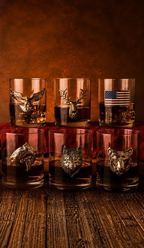 Six clear whiskey glasses arranged on a wooden surface against a warm brown background. Each glass contains amber liquid and has a different metallic animal or flag emblem on the front: eagle, deer, American flag, shark, wolf, and bear.